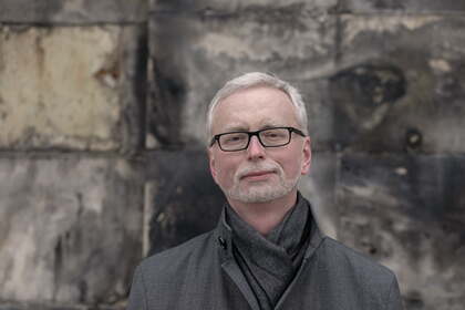 En person med grått hår står framför en vägg med slitna stenar, klädd i grå kappa och halsduk.