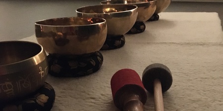 Fem tibetanska klangskålar står på rad på en matta med två klubbor framför dem i ett dämpat rum.