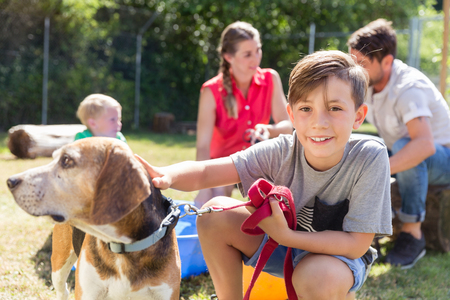En pojke klappar en hund utomhus medan en familj sitter i bakgrunden på en solig dag.