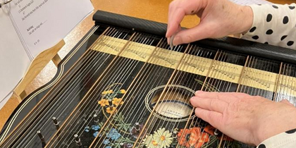 Två händer spelar på en cittra med blommönster, noter syns i bakgrunden.