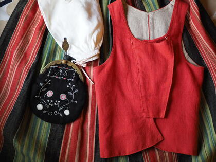 Traditionella klädesplagg inklusive en röd väst, randig kjol, vit mössa och broderad väska ligger utlagda.