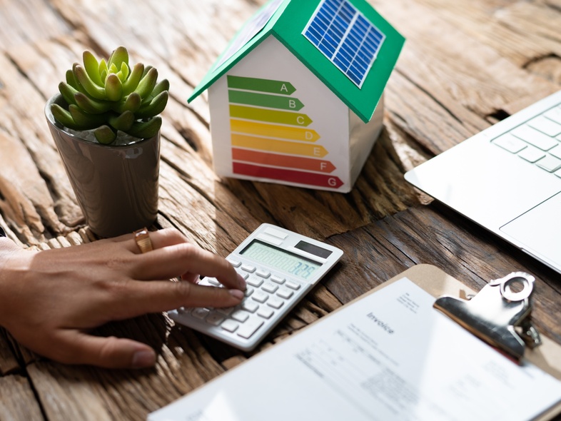 En person använder miniräknare vid ett skrivbord med laptop, pappersarbete, växt och modellhus med energimärkning.