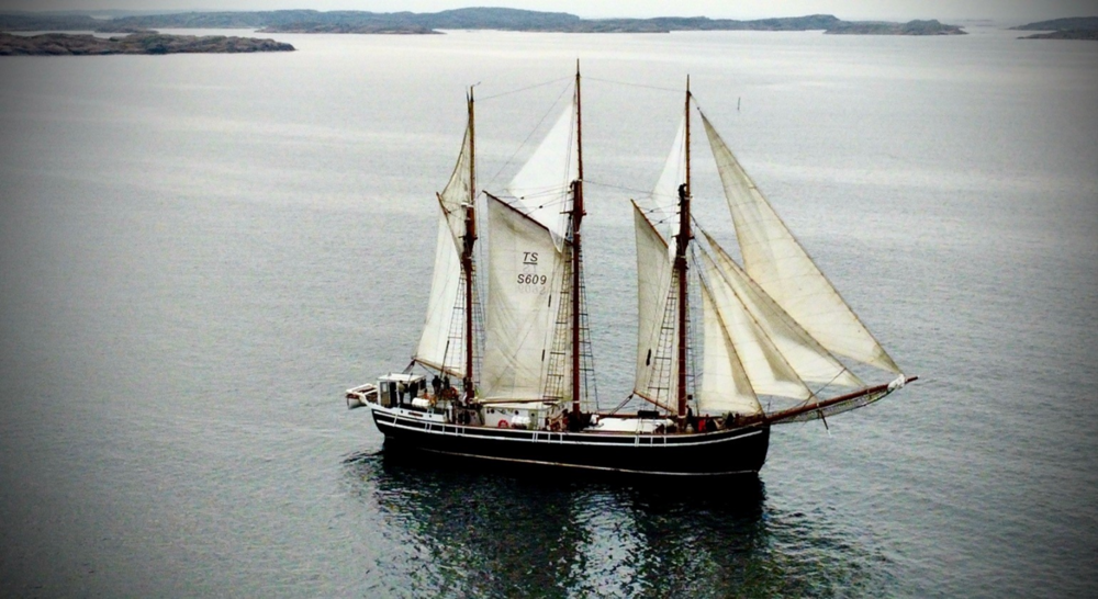 Segelfartyg med tre master seglar på lugnt vatten med öar i bakgrunden.