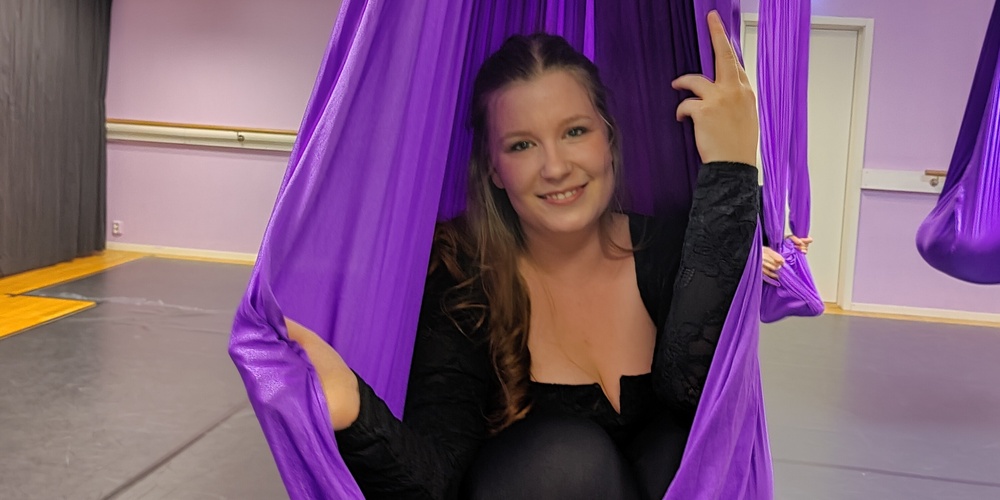 Kvinna sitter i en lila aerial yoga-hängmatta i en träningssal och ler mot kameran.
