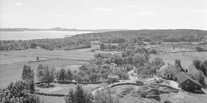 Svartvit landskapsbild över gårdar, åkrar och skog vid en sjö, med slingrande väg och stenmurar.