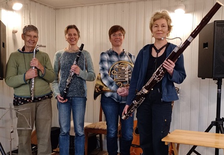 Fyra kvinnor står i ett rum med träpanel och håller olika blåsinstrument, inklusive tvärflöjt, klarinett, valthorn och fagott.