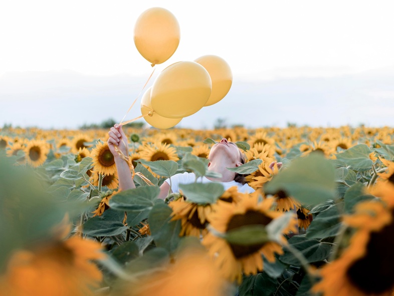 En person håller tre gula ballonger i ett fält av solrosor under en ljus himmel.