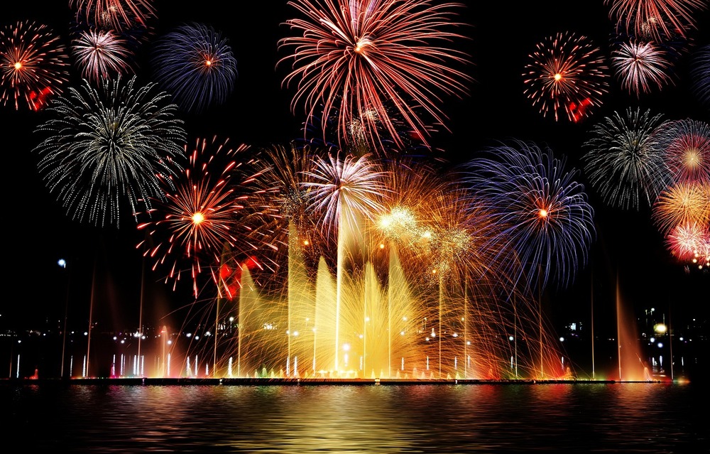 Färgglada fyrverkerier och fontäner lyser upp natthimlen över en sjö.