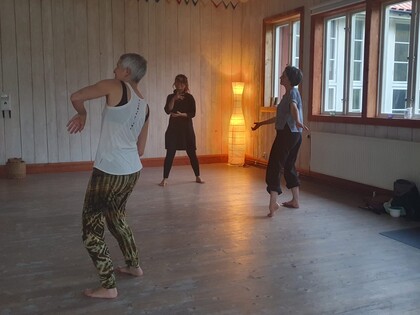 Tre personer dansar barfota i ett ljust rum med trägolv och stora fönster.