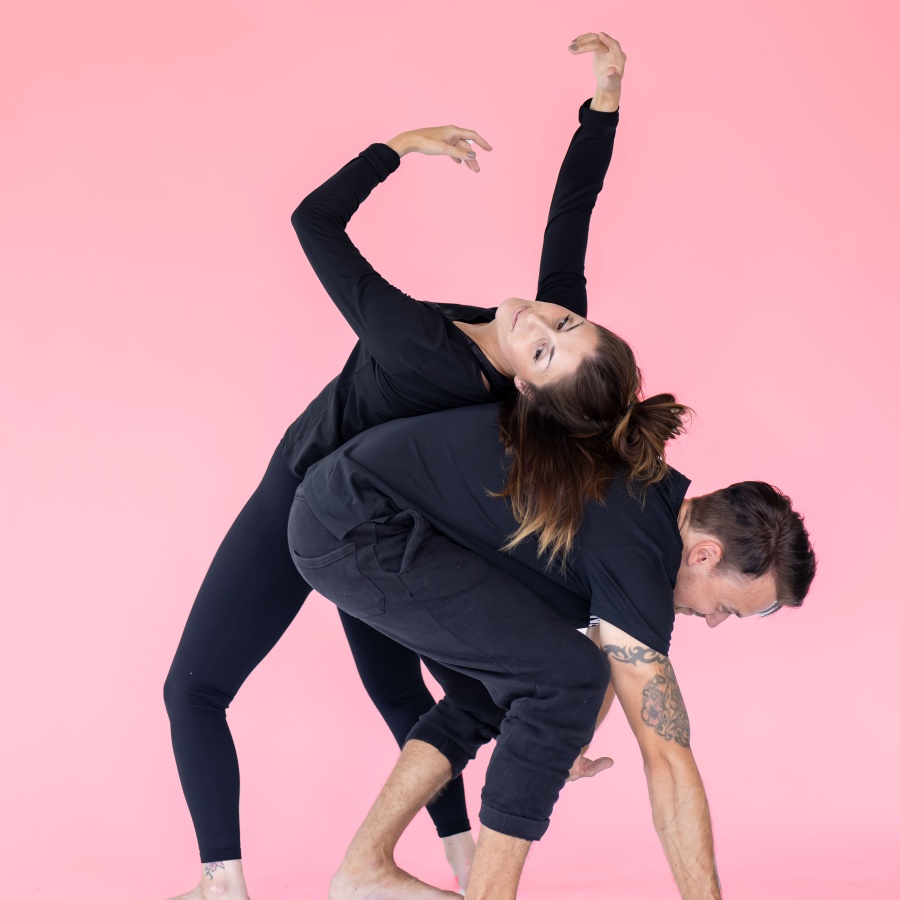Två personer i svarta kläder utför en modern danspose mot en rosa bakgrund.