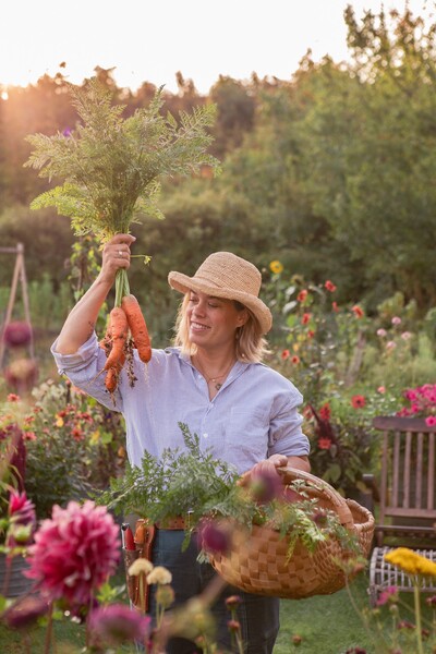 Person i trädgård håller upp nyupptagna morötter och har en korg med grönsaker bland blommor.