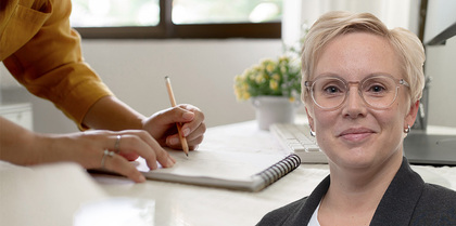 Kvinna med glasögon ler mot kameran, i bakgrunden skriver någon i ett block på ett kontor.
