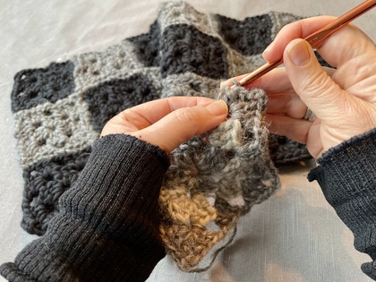 Närbild av händer som virkar med grått garn och virknål, stickad filt i bakgrunden.