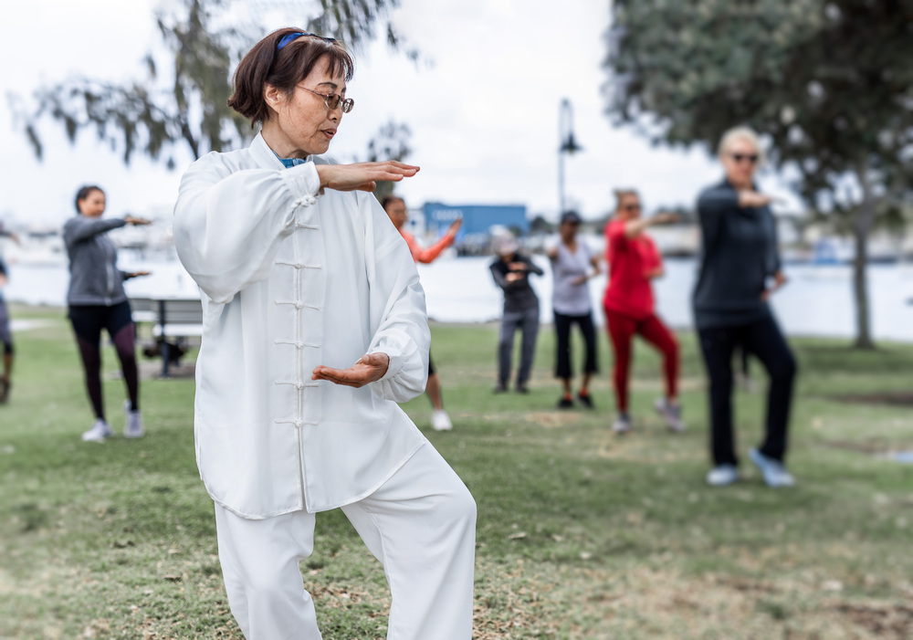 En grupp människor utövar tai chi utomhus i en park, med en kvinna i vit dräkt i förgrunden.