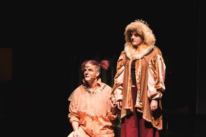 Två personer i teaterkostymer står på en mörk scen, en sitter på knä och den andra står bredvid.