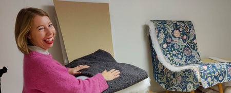Kvinna klär om en stol i ett verkstadsrum, ler mot kameran, arbetsbord med verktyg och tyg syns.