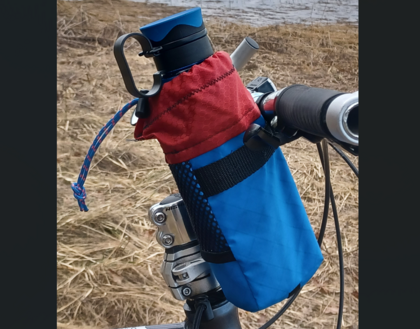 Vattenflaska i blå och röd hållare fäst på ett cykelstyre utomhus vid torrt gräs.