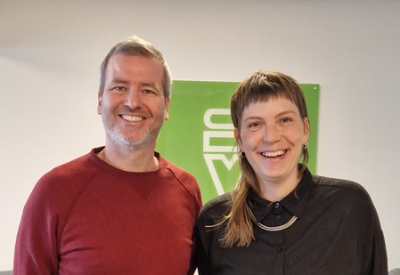 Två leende personer står bredvid varandra inomhus framför en grön skylt.