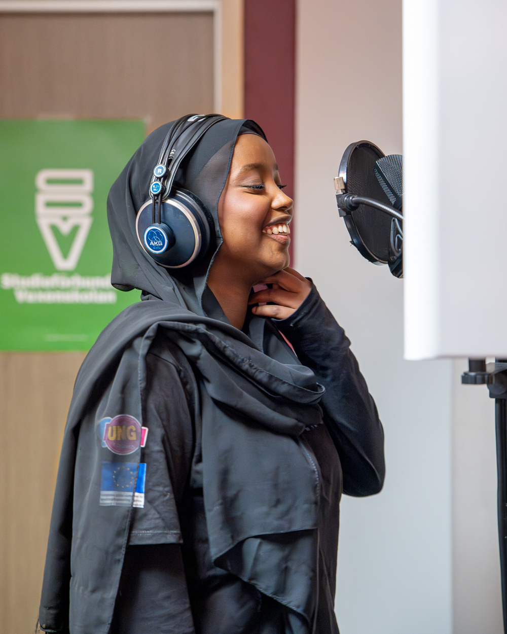 Ung kvinna med hijab och hörlurar sjunger i en mikrofon i en studio.