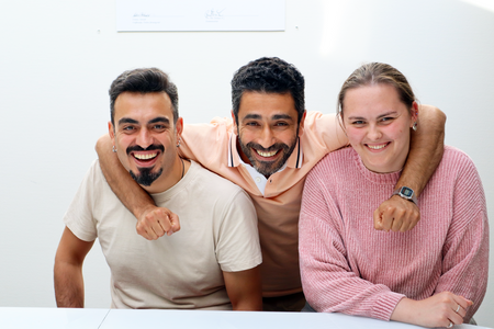 Tre personer sitter tillsammans och ler mot kameran, en man i mitten har armen runt de andra två.