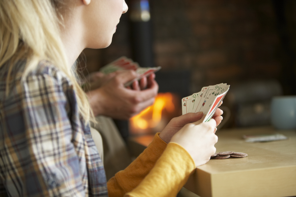 Två personer spelar kort vid ett bord, fokus på händer och kort, med en brasa i bakgrunden.