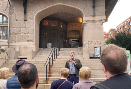 En man talar i mikrofon inför en liten publik utanför en byggnad med stentrappa och stora dörrar.