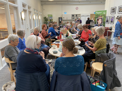 En grupp äldre kvinnor sitter runt bord och handarbetar i en ljus samlingslokal.