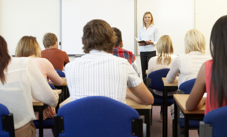 En lärare står framför en klass och undervisar elever som sitter vid bänkar i ett klassrum.