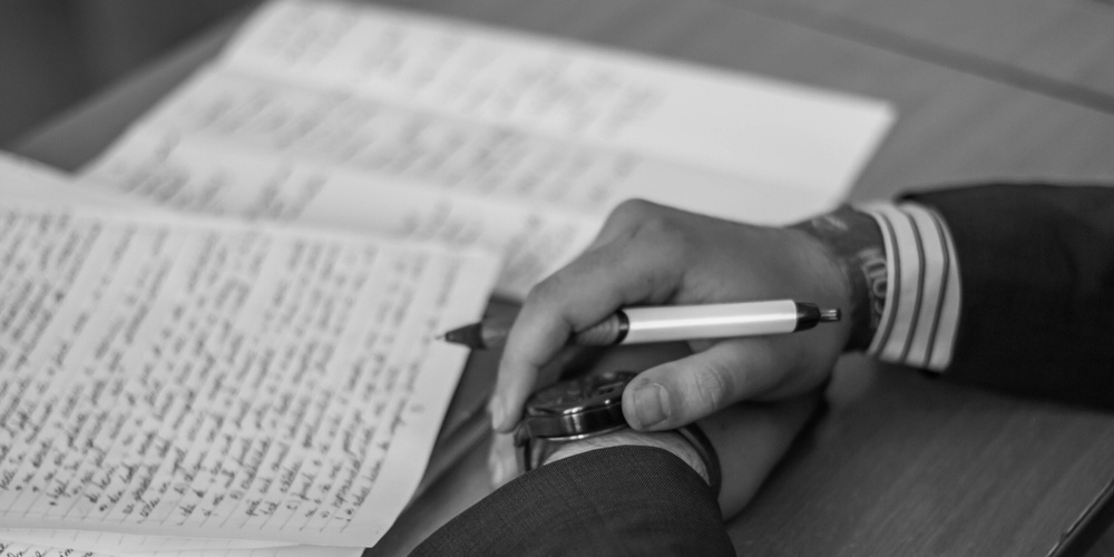 En person i kostym skriver anteckningar för hand på papper vid ett skrivbord, svartvit bild.