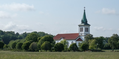 En vit kyrka med rött tak och grön spira står omgiven av träd på en äng under blå himmel.