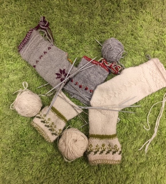 Två par stickade sockor under arbete med garnnystan och stickor på en grön matta.