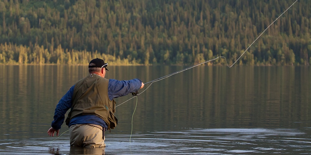 En person står i vattnet och flugfiskar vid en sjö med skogsklädda berg i bakgrunden.
