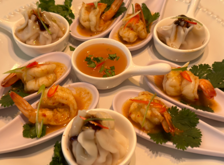 Små skedar med räkor, dumplings och koriander runt en skål med sås på ett vitt fat.