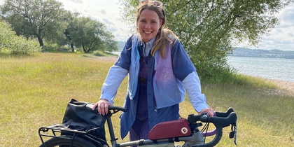 Kvinna står utomhus vid en cykel på en gräsäng nära vatten, med träd i bakgrunden och soligt väder.
