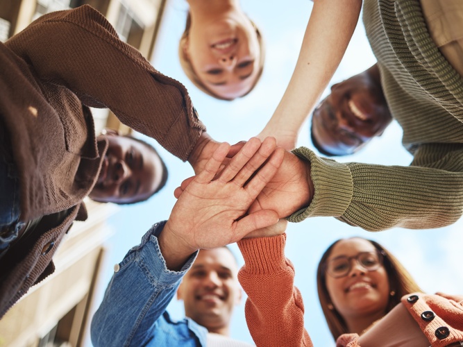 Fem personer står i en cirkel och lägger sina händer ovanpå varandra, sedda underifrån mot himlen.