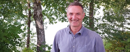 Man i blå skjorta står utomhus framför gröna träd och buskar, ler mot kameran.