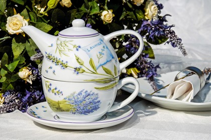 En tekanna och kopp med lavendelmotiv står på ett fat, omgiven av blommor och en servett på tallrik.