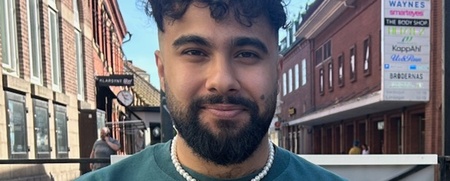 Man med lockigt hår och skägg sitter utomhus vid ett bord med pizza och dryck framför sig.