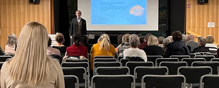 En föreläsare står framför en publik i en sal med träpaneler och visar en presentation på en stor skärm.