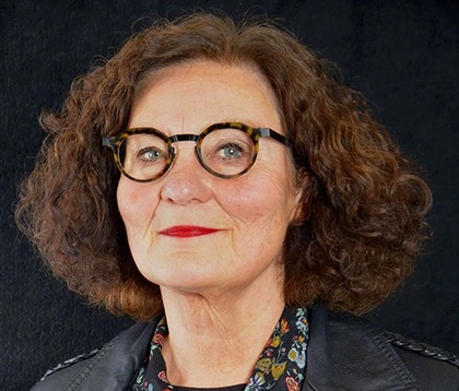 Person med lockigt brunt hår, svart läderjacka och blommig skjorta mot svart bakgrund.