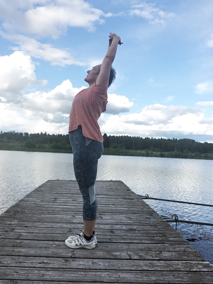 Person står på en brygga vid en sjö och sträcker armarna mot himlen under en blå himmel med moln.