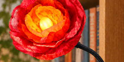 En blomformad lampa med klämfäste sitter fast på en bokhylla, omgiven av böcker och växter.