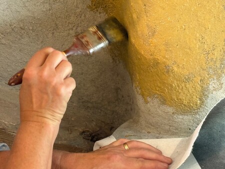 En hand målar en ojämn vägg med gul färg med hjälp av en pensel.