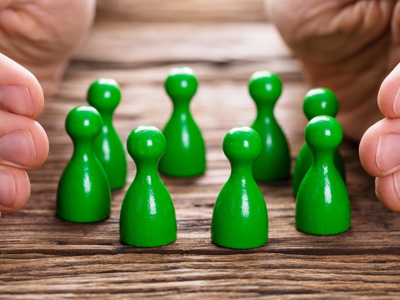 Två händer skyddar en grupp gröna spelpjäser på ett träbord.