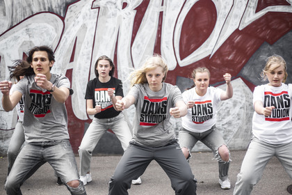 Fem unga personer dansar energiskt framför en graffitivägg, alla bär t-shirts med texten 'Dansstudio'.