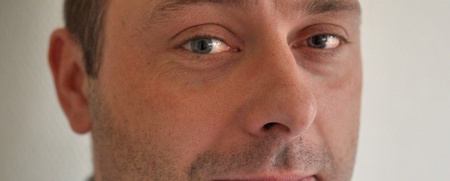 Man med kort brunt hår och ljusblå skjorta tittar in i kameran mot en ljus bakgrund.