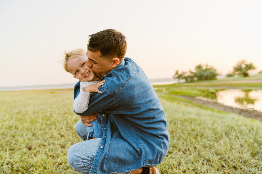 En vuxen och ett barn kramas och skrattar utomhus på en grön äng vid solnedgången.