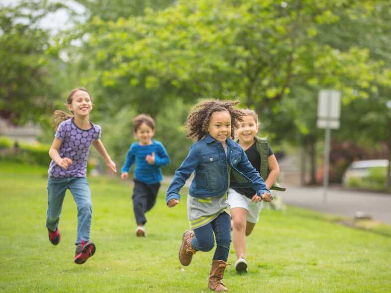 Fyra barn springer över en gräsmatta med gröna träd i bakgrunden.