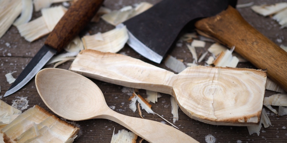 En träsked, en yxa och en kniv ligger på ett bord omgivet av träspån och en halvfärdig träsked.