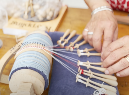 Närbild av händer som tillverkar spets med hjälp av knyppelpinnar och en knyppeldyna.